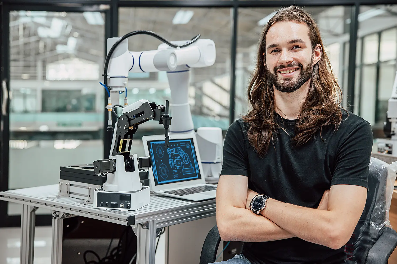 A young man is sitting at a workbench with a robotic arm and a laptop on it.